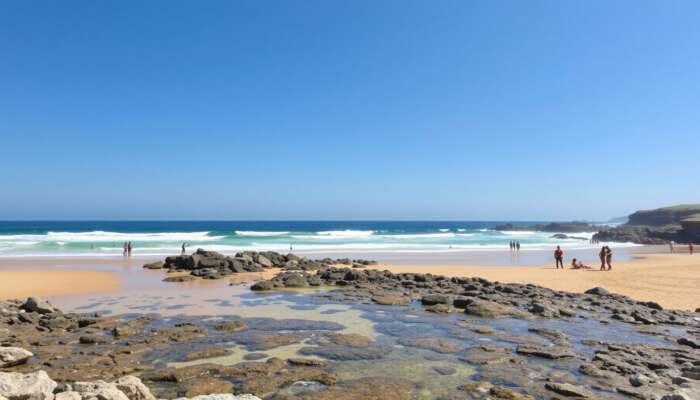 A sunny beach featuring clear waves, golden sands for sunbathing, rocky outcrops for exploration, accessible ramps, and diverse people enjoying the day under a blue sky.
